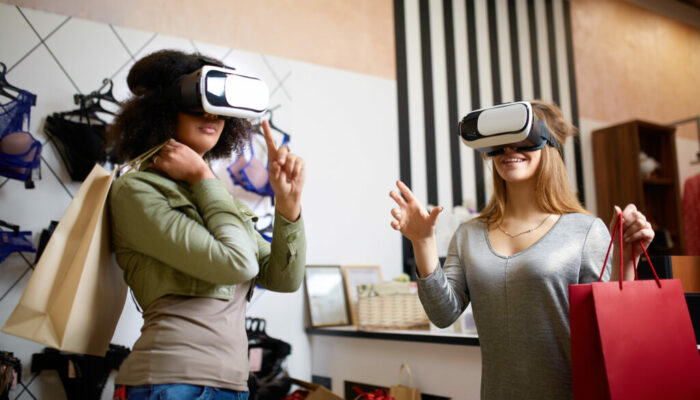 Technological concept. Two women in modern virtual reality headsets having expirience in shopping at lingerie store. Multiracial girls in vr glasses with bags touching and pointing interface elements in underwear shop.