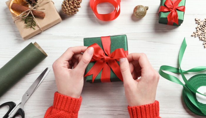 Christmas present. Woman tying ribbon bow on gift box at white wooden table, top view